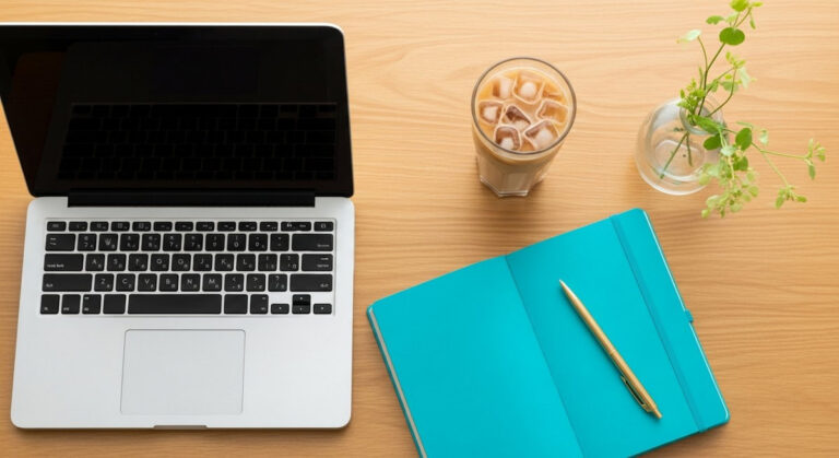 a flatlay of a laptop and teal notebook on a wooden desk ready to work from home for selling on Etsy