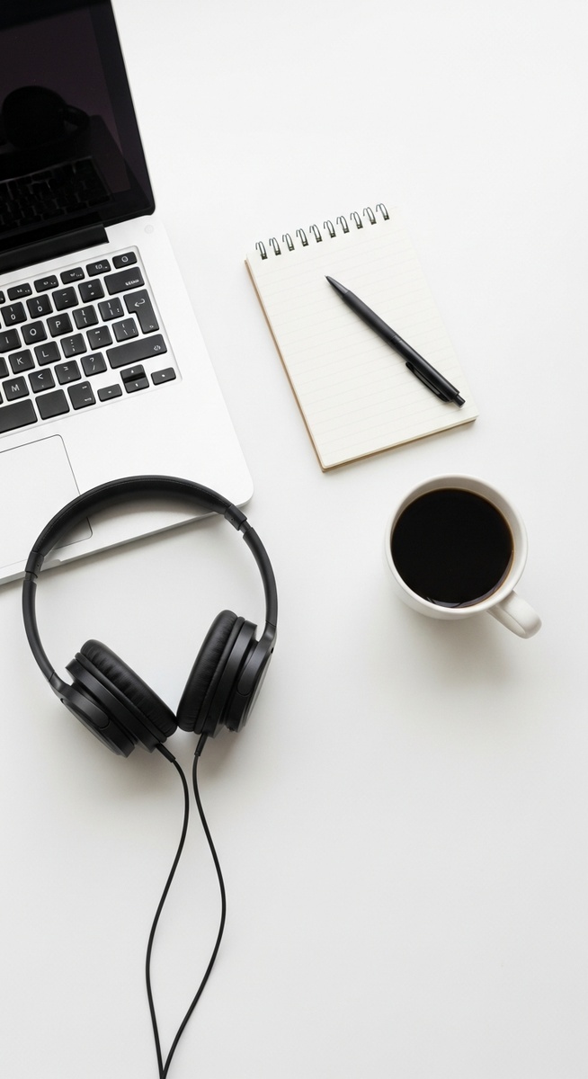 a flatlay of a laptop, headphones, notepad and coffee mug on a white desk to work from home